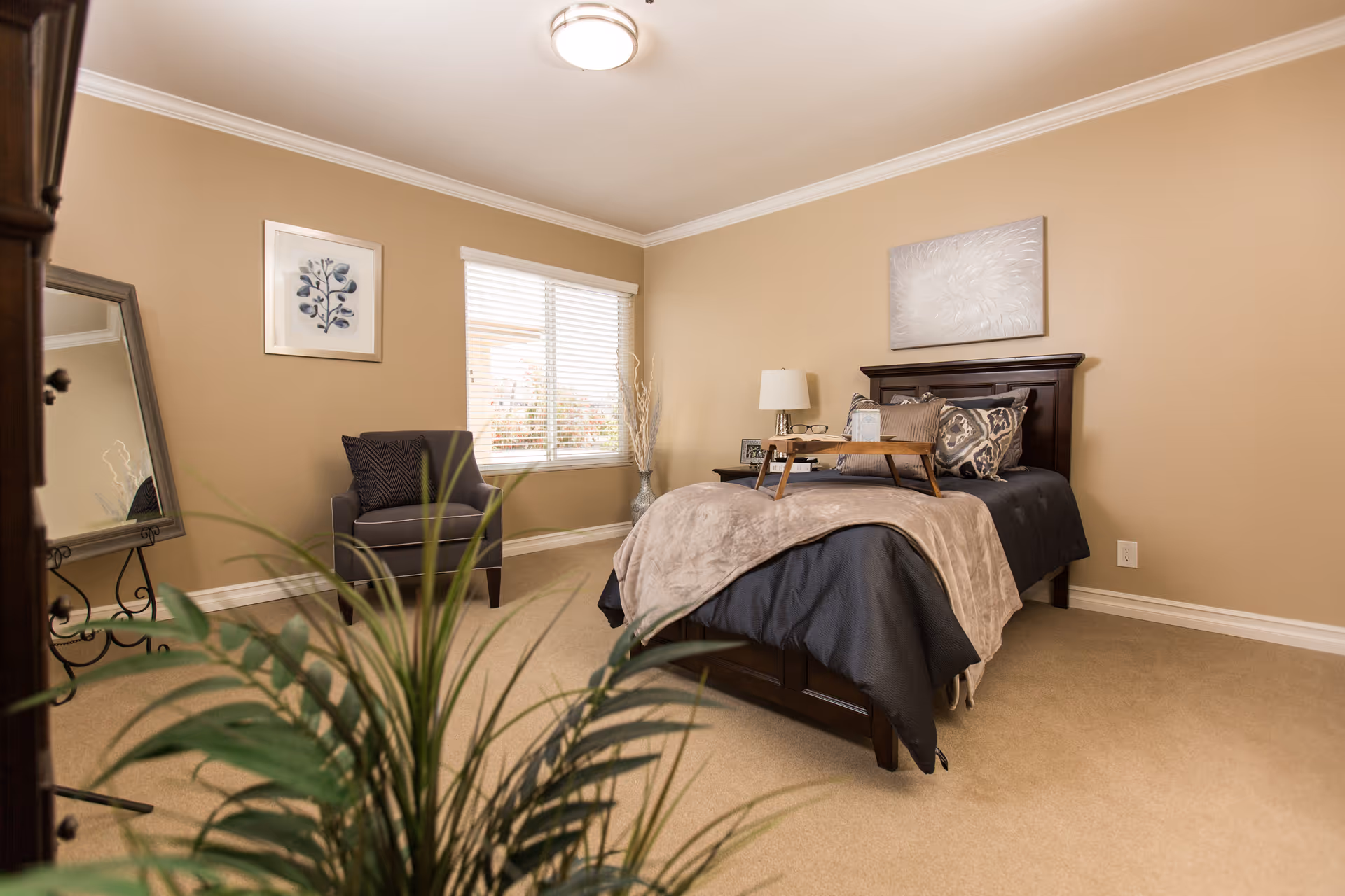 Neat, softly lit bedroom with a made bed, armchair, window, and decorative plants.