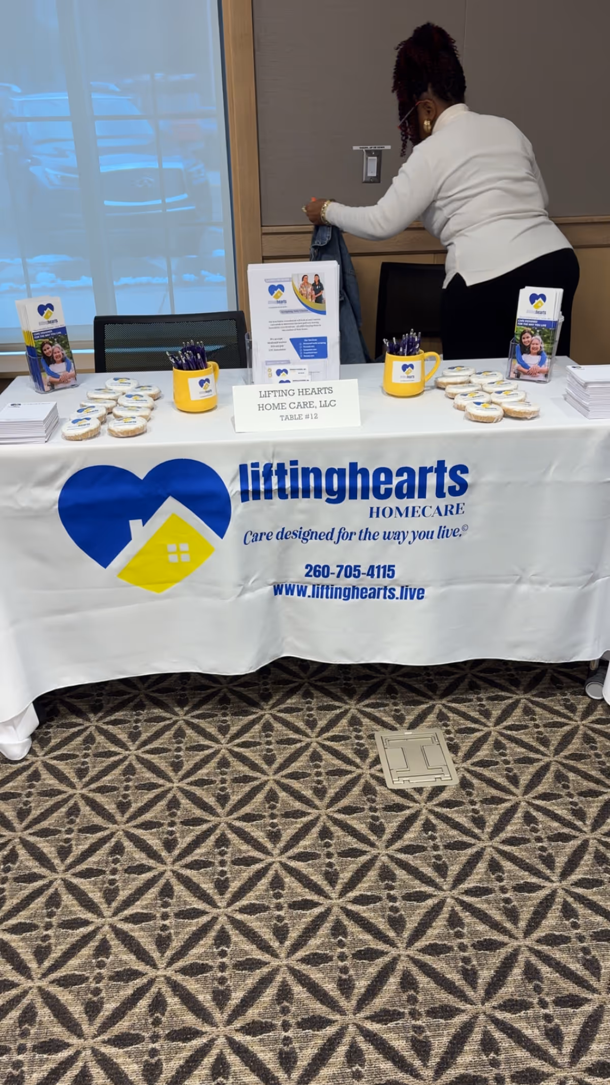 A staff member arranges a Lifting Hearts Home Care promotional table indoors.