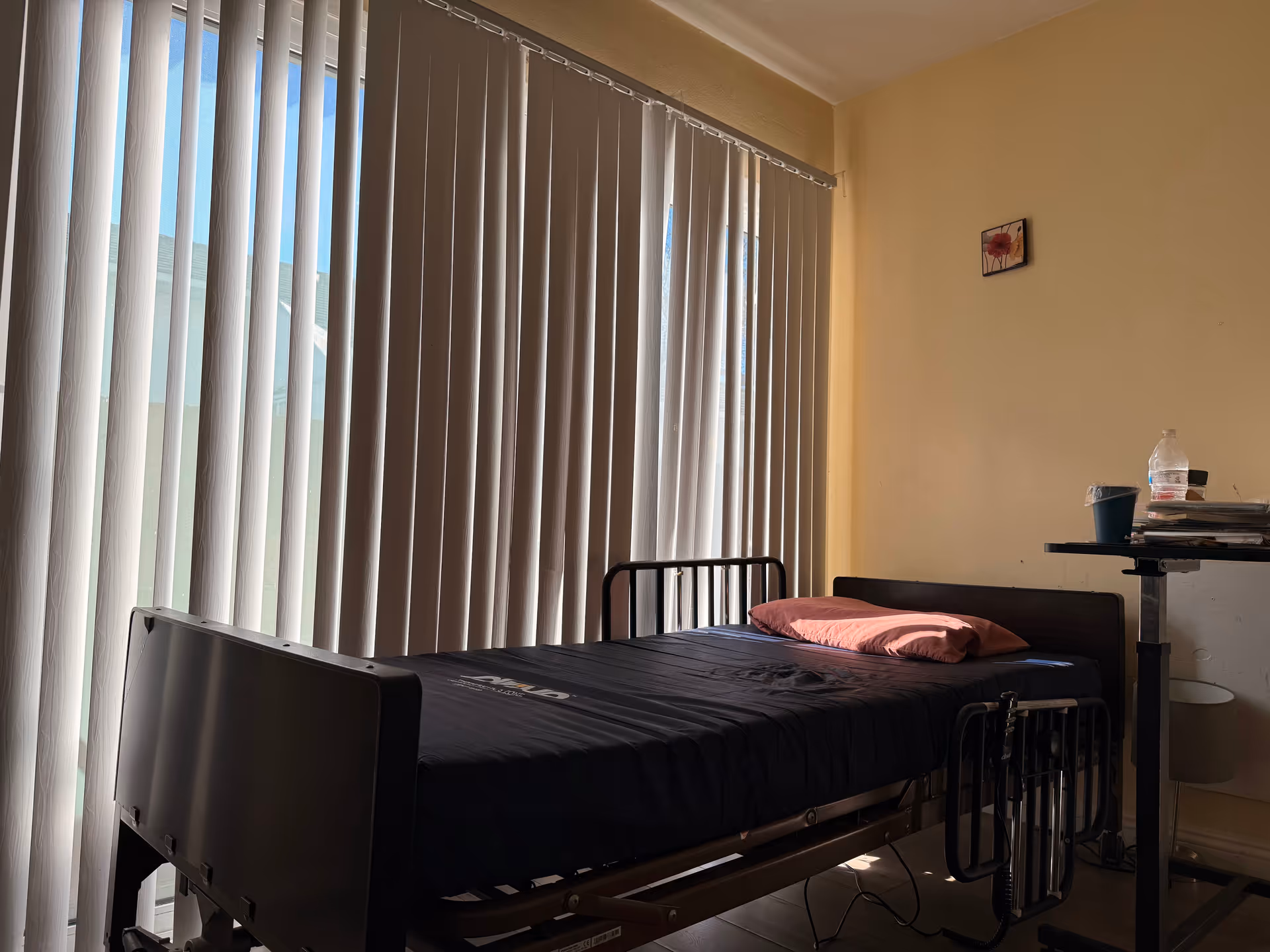 A single hospital-style bed in a seniors' room with vertical blinds and a small table.