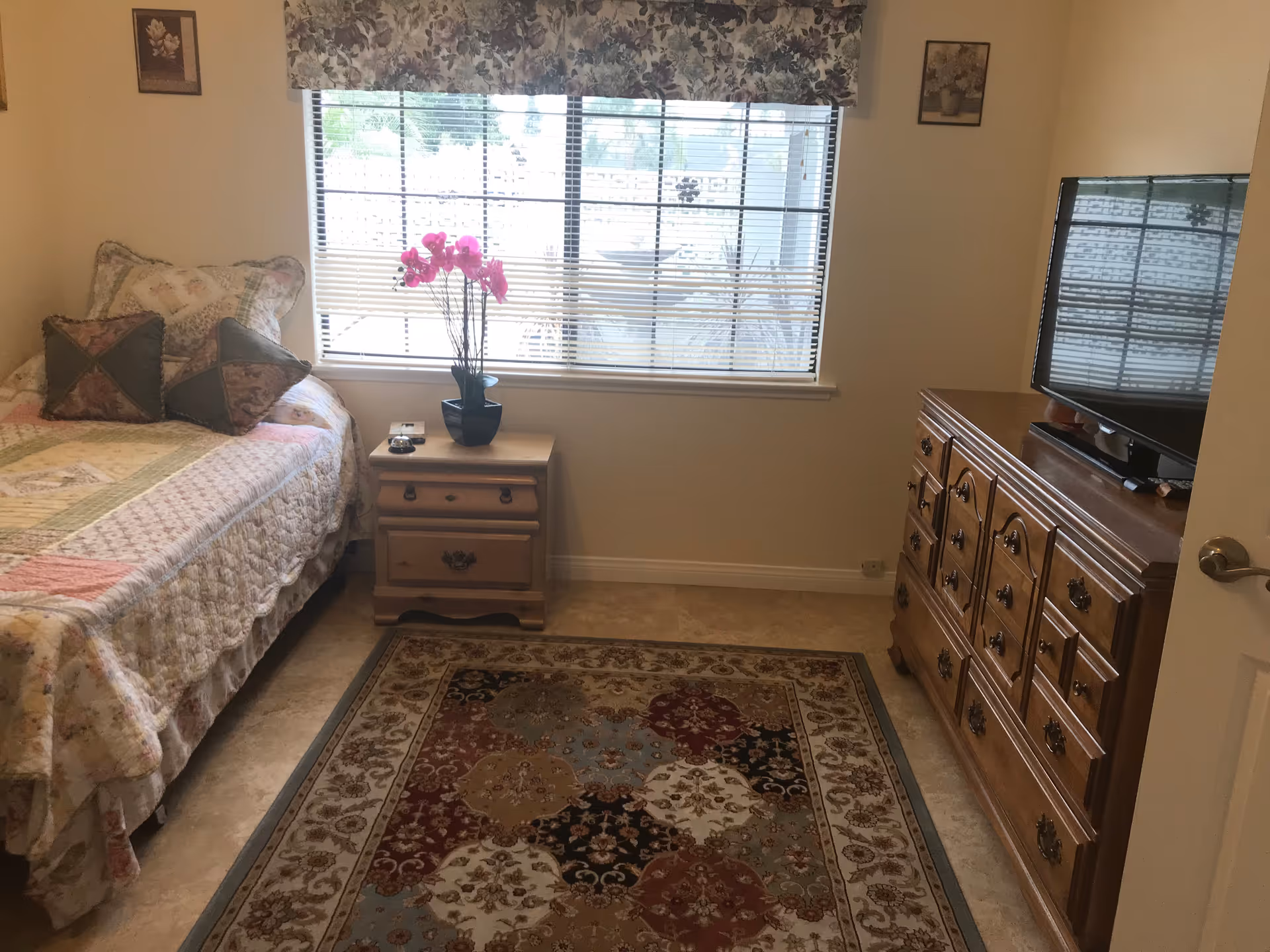 A cozy bedroom with a bed, nightstand, dresser, rug, window blinds, and a TV.