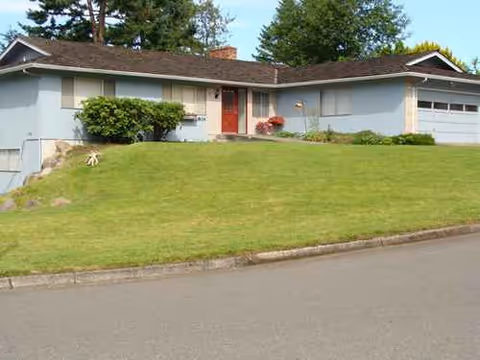 Front view of a single-story blue house with a red door, shrubs, and a driveway.