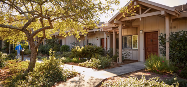 Sunlit landscaped walkway in front of single-story cottage-style units with a person walking at the left.