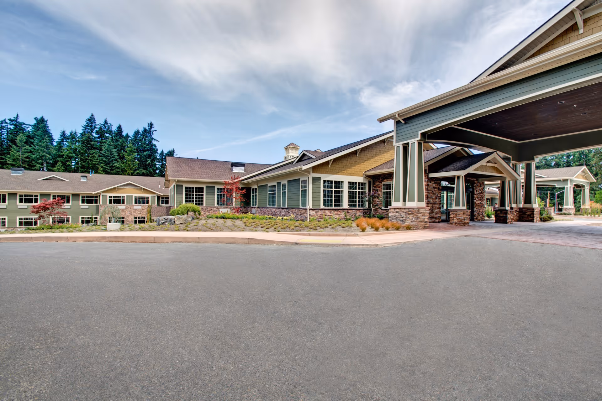 Exterior view of Life Care Center of South Hill showing a large building with multiple windows, a covered entrance supported by stone pillars, and landscaped grounds with trees and shrubs under a partly cloudy sky.