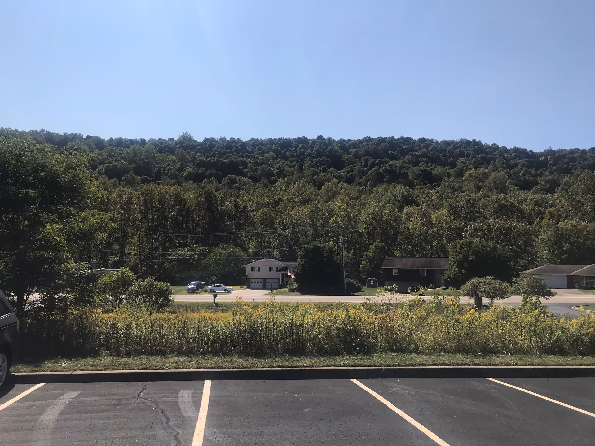 Parking lot in foreground, grassy verge, white building across street, surrounded by trees.