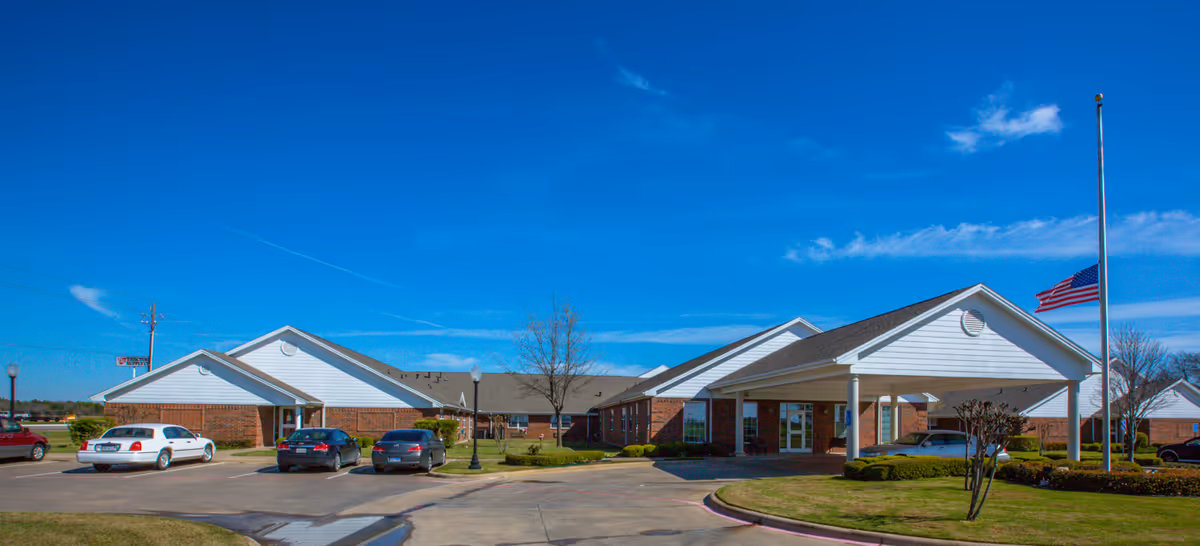 Exterior view of Wesley House senior living facility showing a single-story brick building with white siding and a covered entrance. Several cars are parked in front, and an American flag is flying at half-mast on a flagpole. The sky is clear and blue.