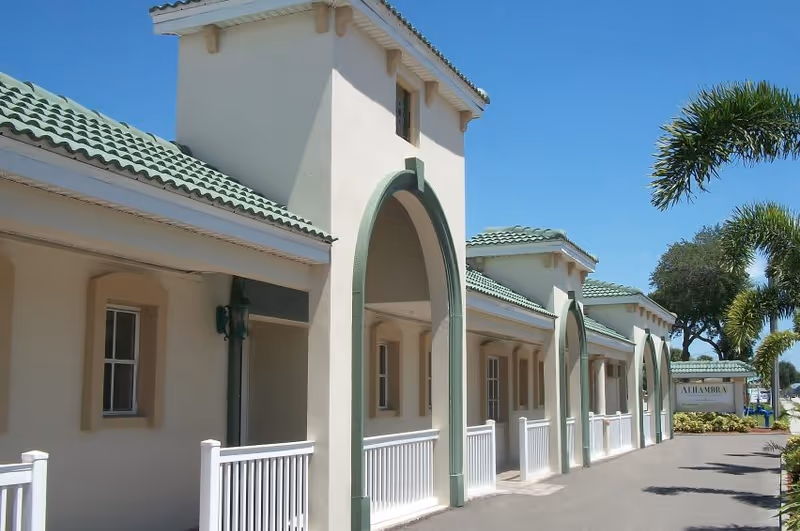 Front exterior of Alhambra Health and Rehabilitation Center featuring a beige building with green-tiled roofs, arched entrances, a white railing, and palm trees.
