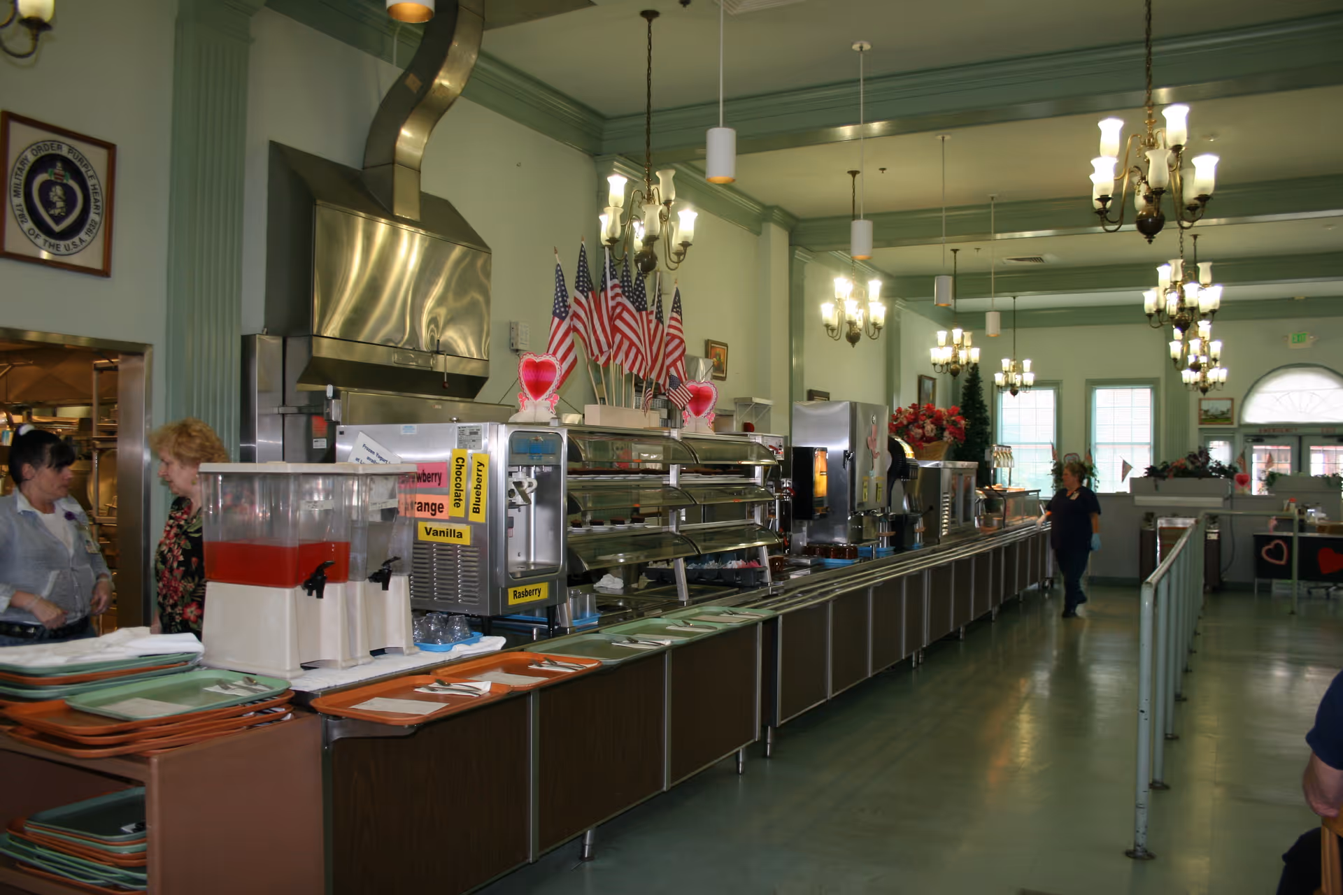 Cafeteria-style dining room with a long serving line, beverage dispensers, American flags, chandeliers, and a few people.