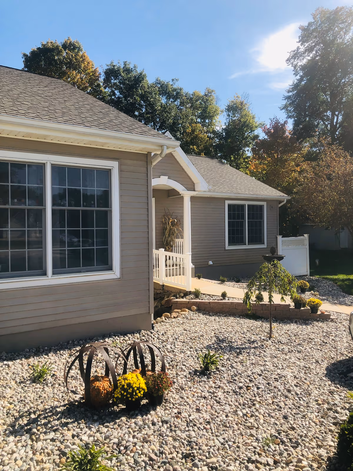 Exterior front of a single-story beige building with a small white porch, rock-covered yard with potted flowers and decorative metal accents.