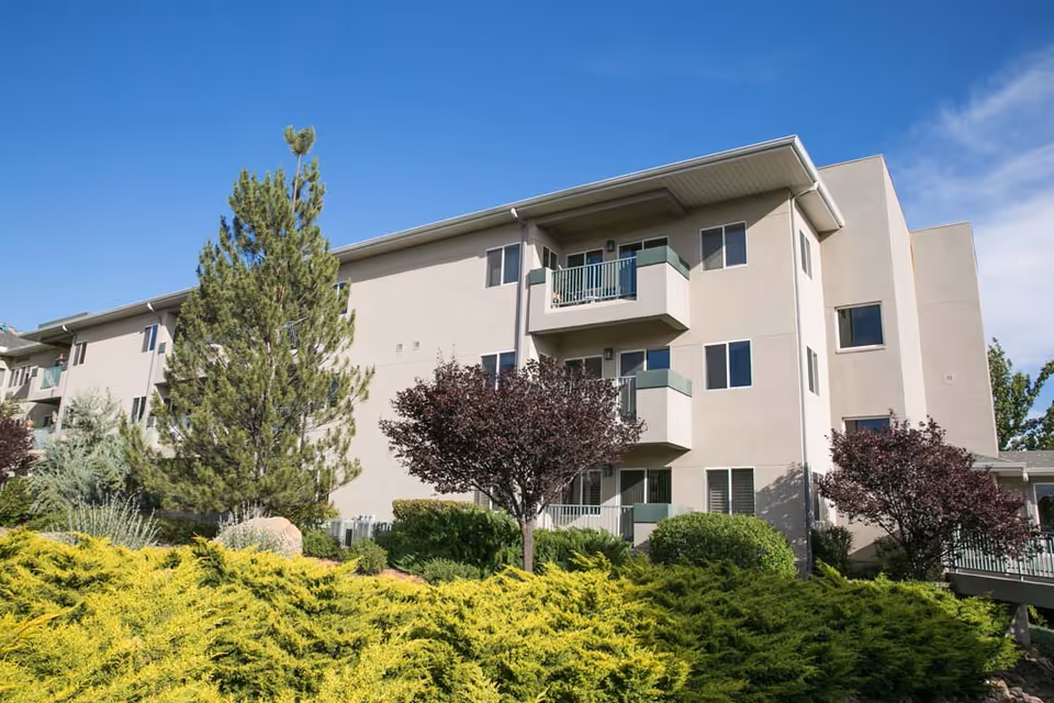 Exterior view of a three-story beige residential building with balconies, surrounded by green shrubs, trees, and a clear blue sky.