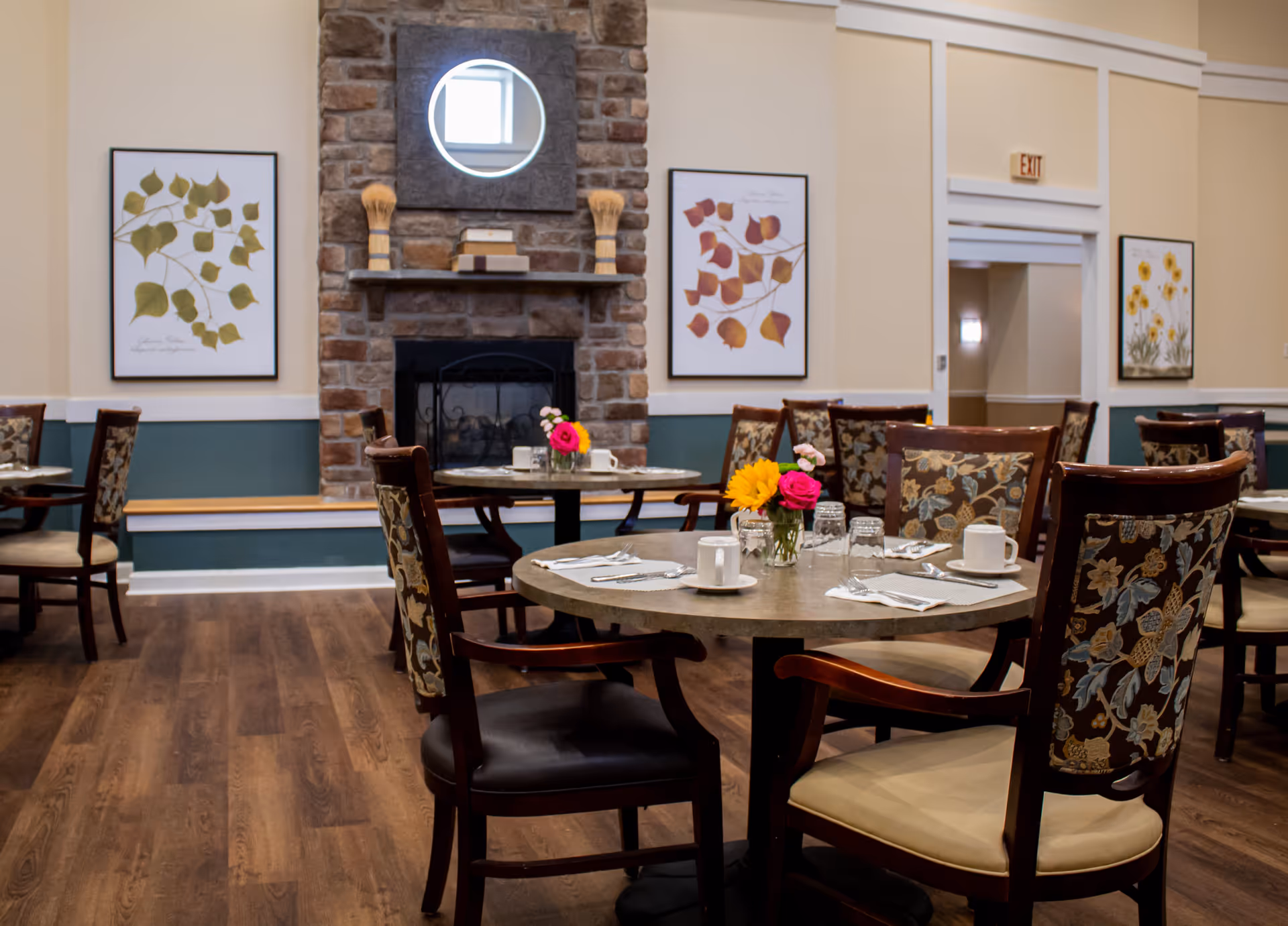 A cozy dining room with round tables set with cups, glasses, and silverware. Each table has a small vase with colorful flowers. The room features wooden chairs with floral upholstery, a stone fireplace with a circular mirror above it, and framed botanical prints on the walls. The floor is wooden, and there is an exit door in the background.
