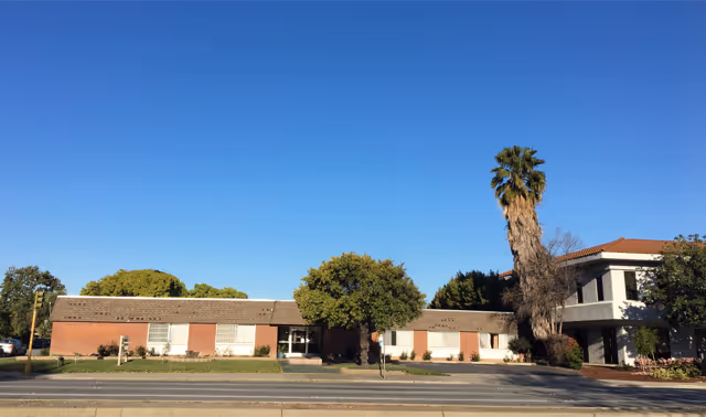 Exterior view of a single-story brick building with a tiled roof, several windows, and a central entrance. There is a tall palm tree and other greenery in front of the building, with a clear blue sky overhead.