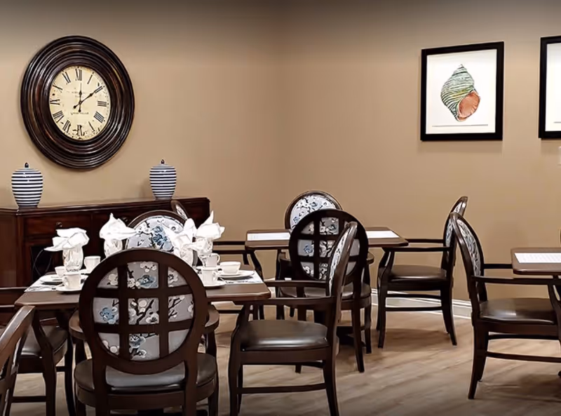 Dining room with several wooden tables and chairs featuring floral patterned backs. Tables are set with white napkins folded in decorative shapes, cups, and plates. A large round wall clock and two striped vases are on a wooden sideboard against a beige wall. Two framed pictures of seashells hang on the wall.