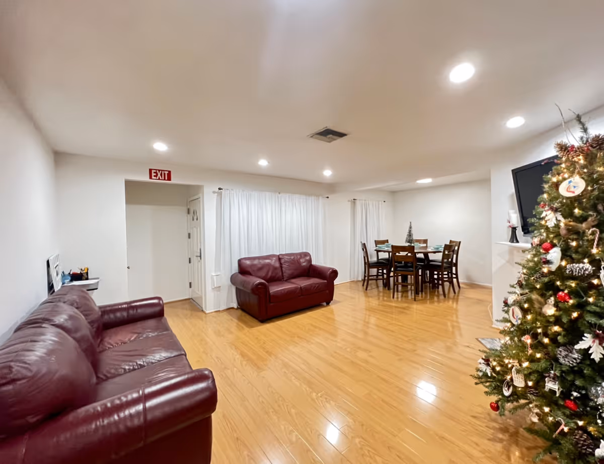 A spacious living room with wooden flooring featuring two red leather sofas, a decorated Christmas tree, a wall-mounted TV, and a dining area with a wooden table and chairs. White curtains cover the windows, and there is an exit sign above a doorway.