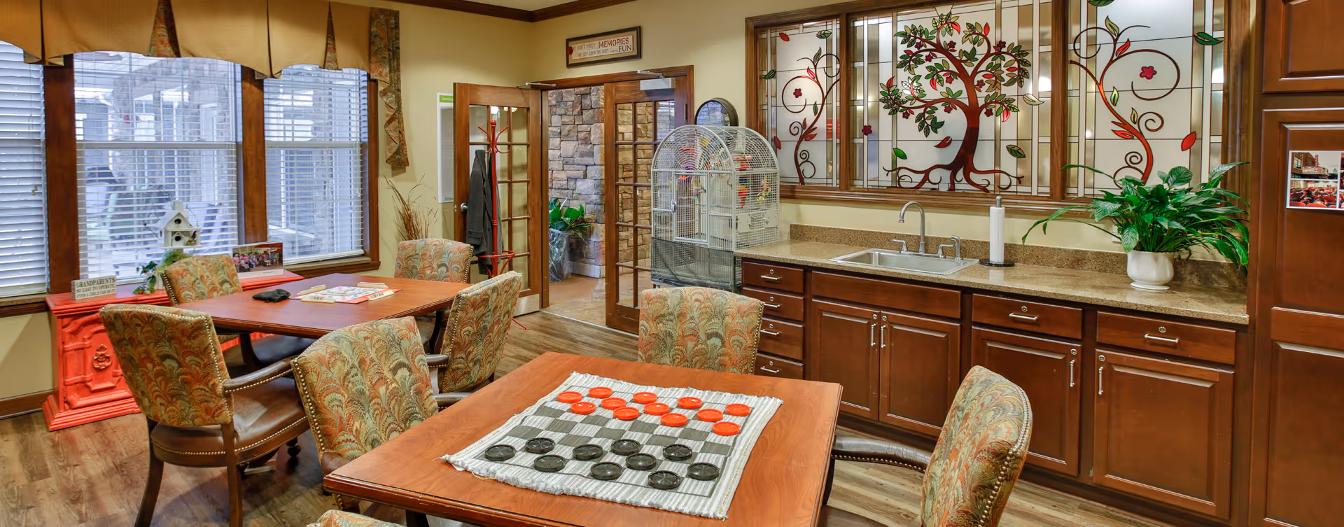 A cozy common area with a wooden table and patterned chairs arranged around it. On the table is a checkers game set up with red and black pieces. Behind the table is a countertop with a sink, a potted plant, and a large birdcage. The wall above the countertop features decorative stained glass panels with tree and leaf designs. There are windows with blinds and curtains on the left side, and a door with glass panels leading to another room. The floor is wooden, and the overall atmosphere is warm and inviting.