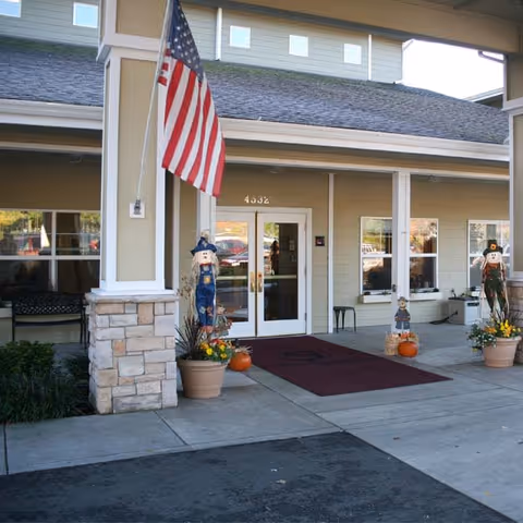Entrance of a building with double glass doors, decorated with two scarecrow figures, potted plants, and pumpkins. An American flag is mounted on a pillar to the left of the entrance. The building has beige siding and stone accents on the pillars.