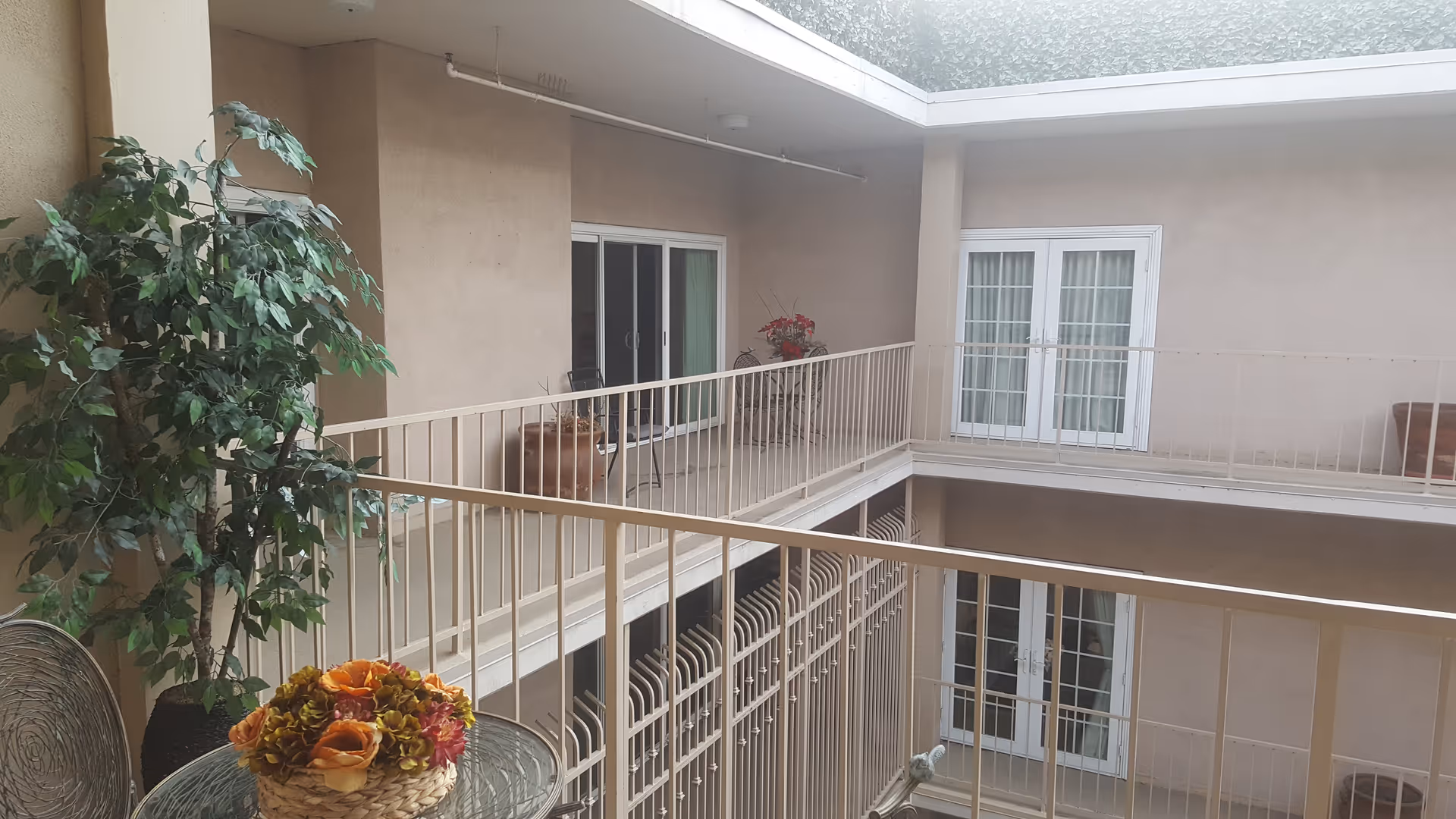 Two-story indoor courtyard atrium with railings, doors, potted plants, and a table with flowers.