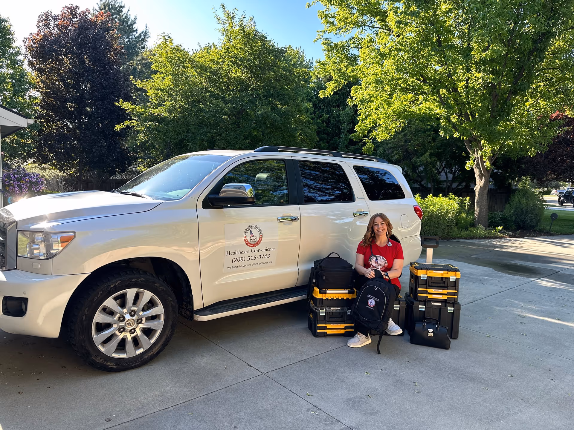 A smiling woman sits beside a white healthcare concierge van with gear on a driveway.