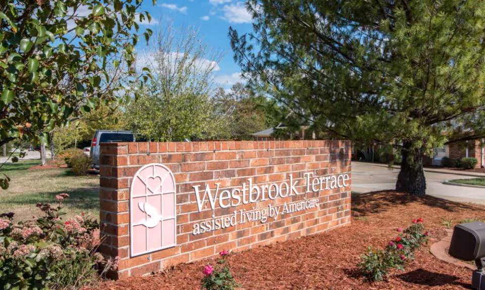 A brick sign for Westbrook Terrace assisted living by Americare, surrounded by landscaped plants and trees under a blue sky with some clouds.