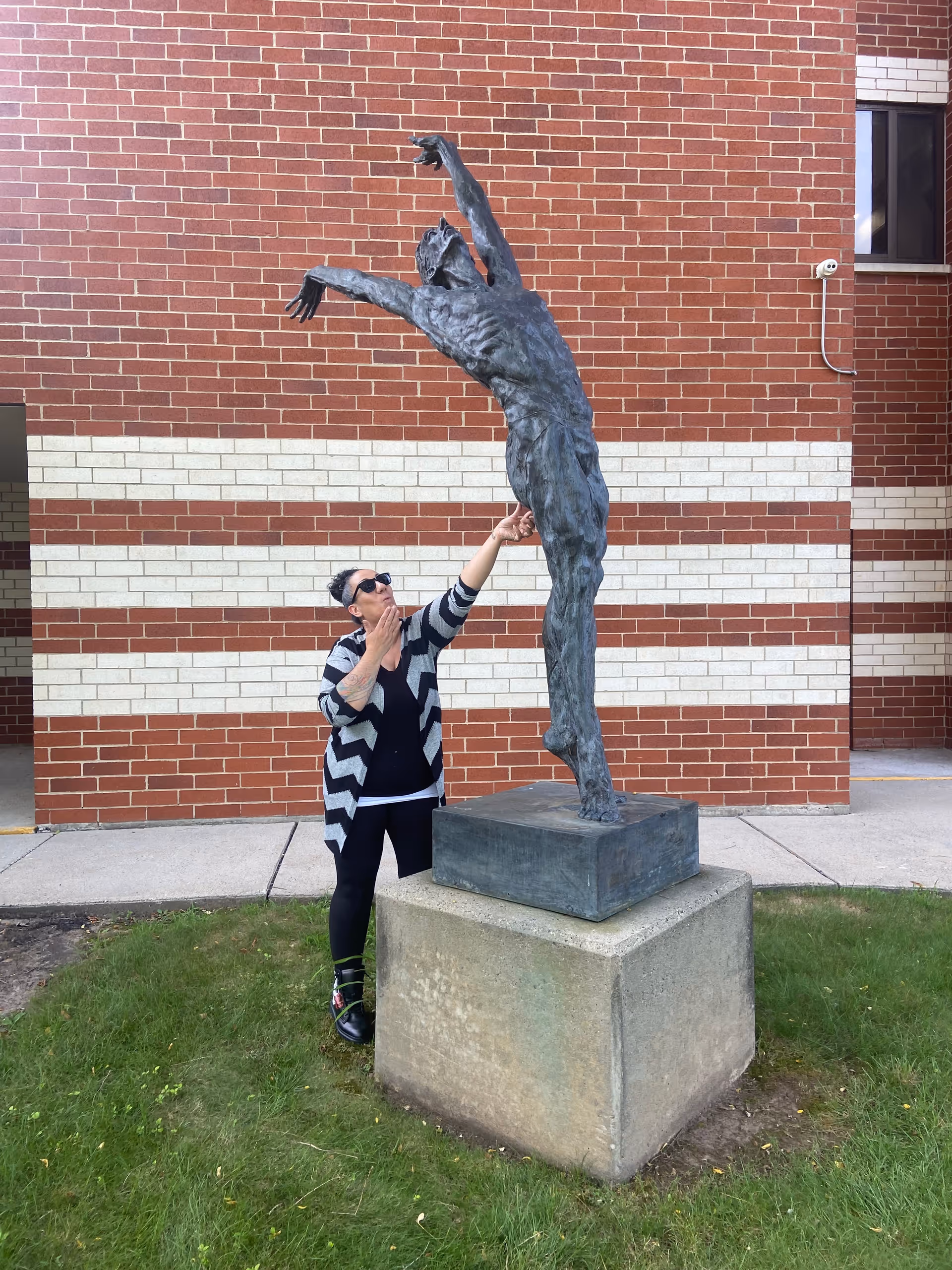 Person wearing sunglasses poses beside a tall statue outside a brick building.