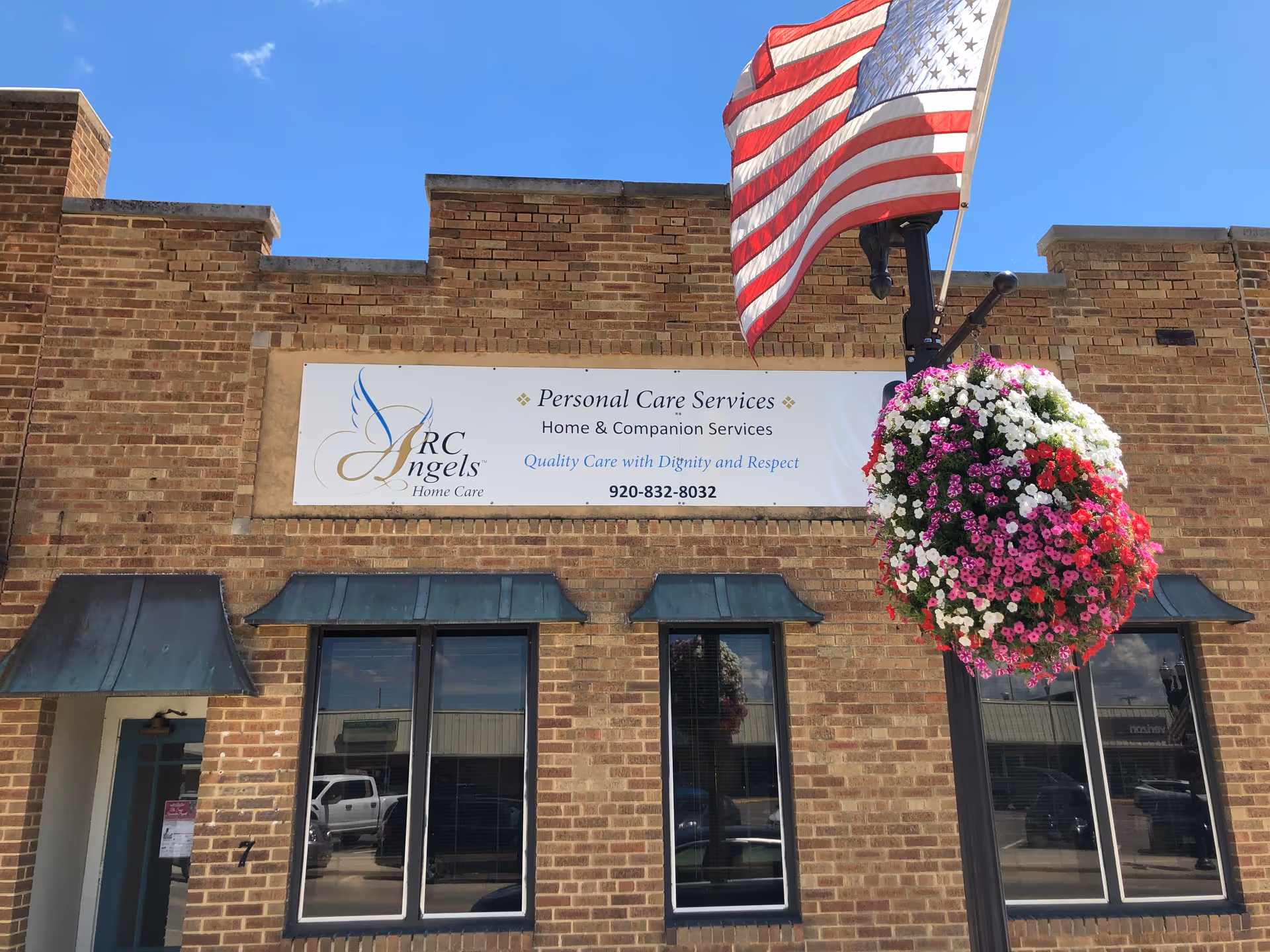 Brick building facade with ARC Angels Home Care sign, American flag, and hanging flower basket.
