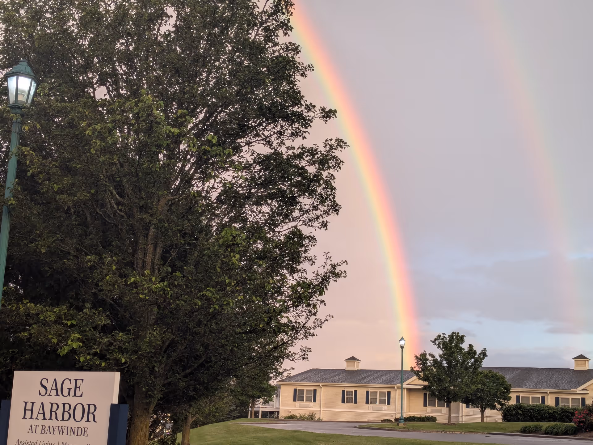 Rainbow arches over Sage Harbor at Baywinde exterior with sign and trees.