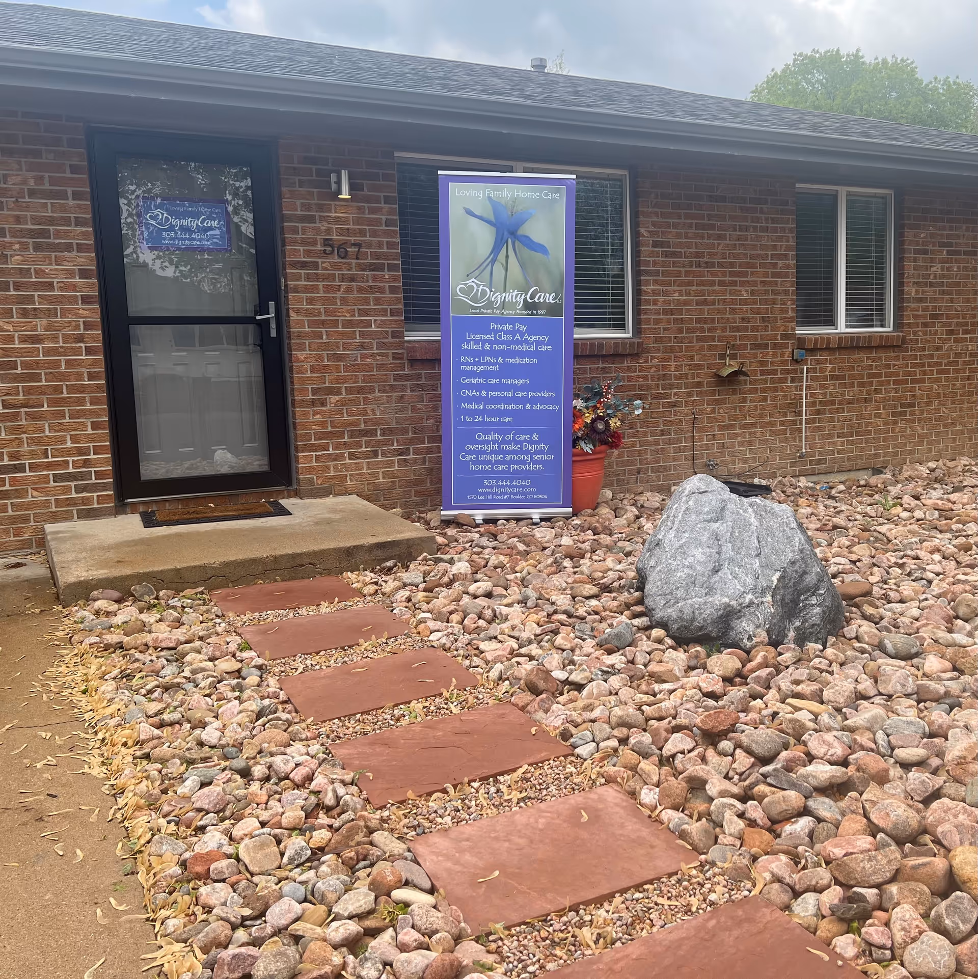 Exterior view of a brick senior living facility entrance with rock garden and a banner sign.