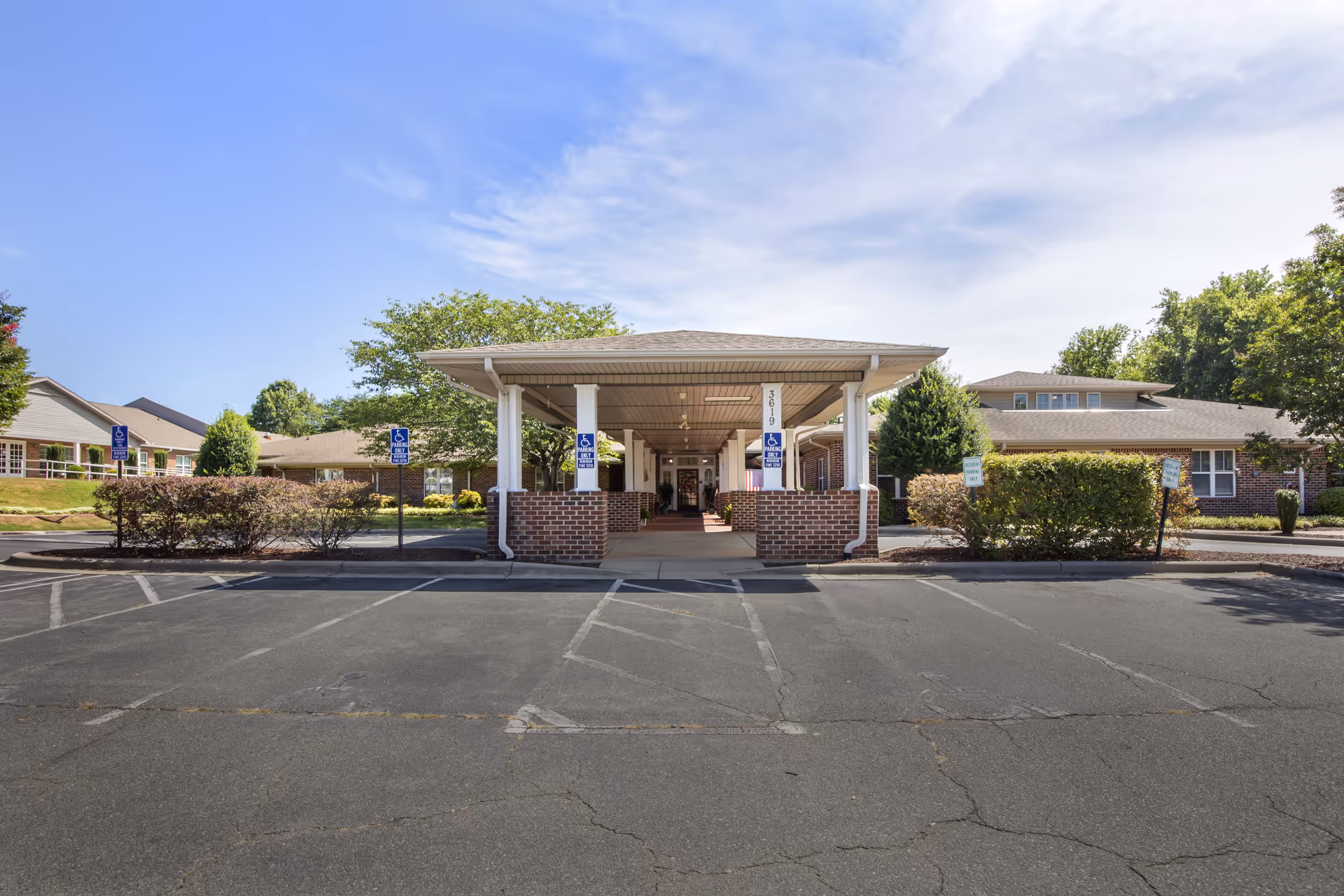 Front entrance of Brookdale Burlington facility with a covered drop-off area supported by white columns and brick bases. The building is surrounded by greenery and parking spaces, including designated handicapped parking spots.