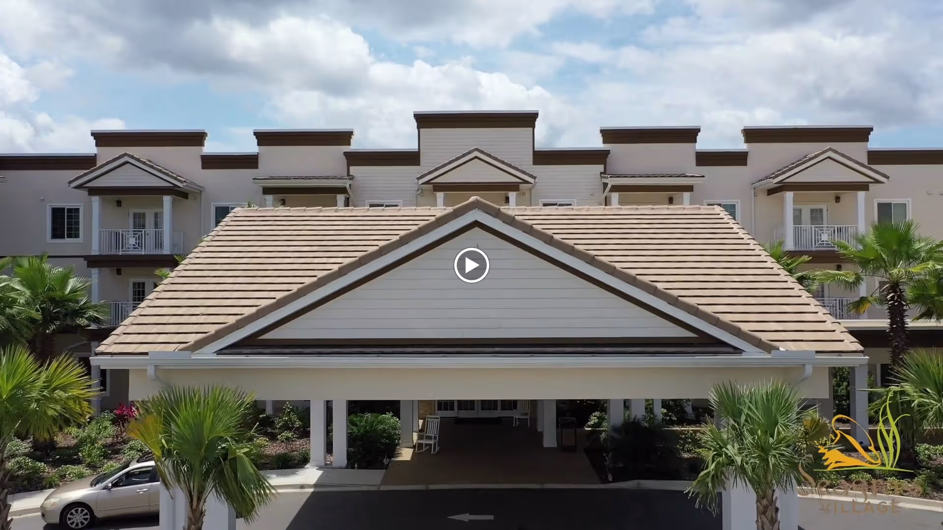 Front exterior view of a multi-story senior living facility building with a covered entrance, palm trees, and landscaped greenery under a partly cloudy sky.