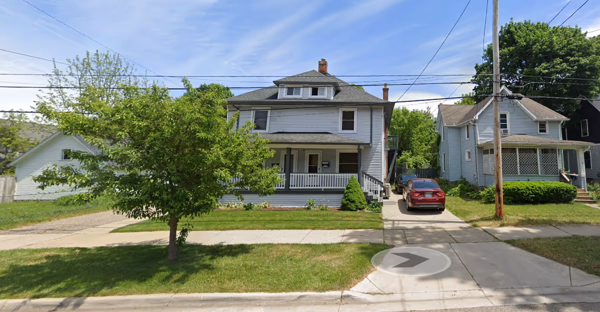Two-story gray house with a front porch, trees, and a driveway along a residential street.
