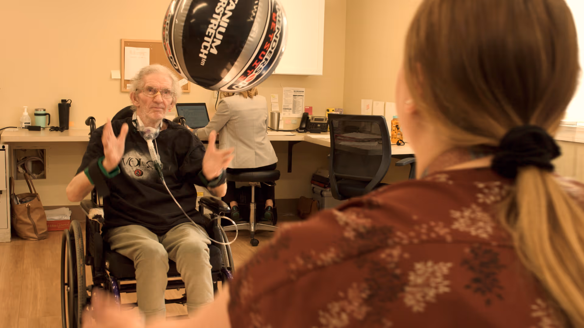 An elderly man in a wheelchair playing with a black and white basketball indoors, while a woman with a ponytail faces him. In the background, another person is seated at a desk working on a computer in a room with beige walls and wooden flooring.
