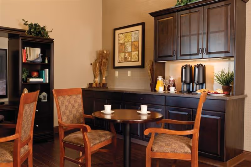 A cozy seating area with a round wooden table and four cushioned wooden chairs. Behind the table is a dark wood cabinet with glass doors on the upper section and solid doors below. On the countertop are beverage dispensers, a pitcher of orange juice, cups, and a small plant. To the left, there is a tall dark wood bookshelf with decorative items and books. The walls are painted beige with a framed artwork hanging above the cabinet.