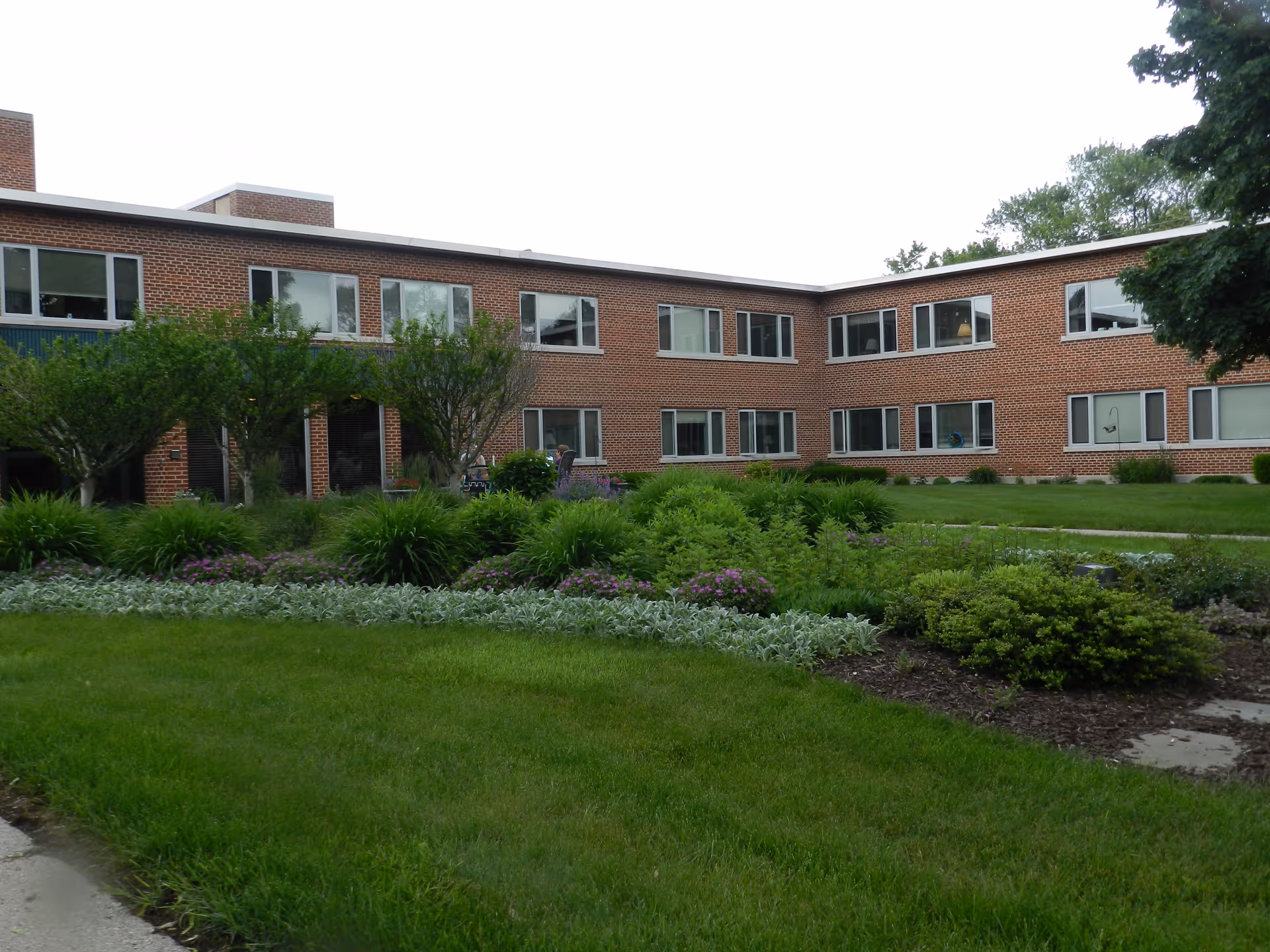 Brick two-story senior living building with landscaped lawn and garden in foreground.