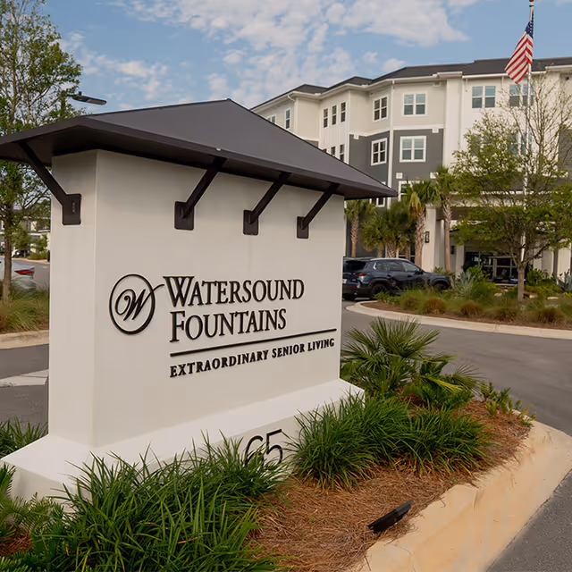 Entrance sign for Watersound Fountains senior living facility with landscaped greenery around it and a multi-story building in the background under a partly cloudy sky.