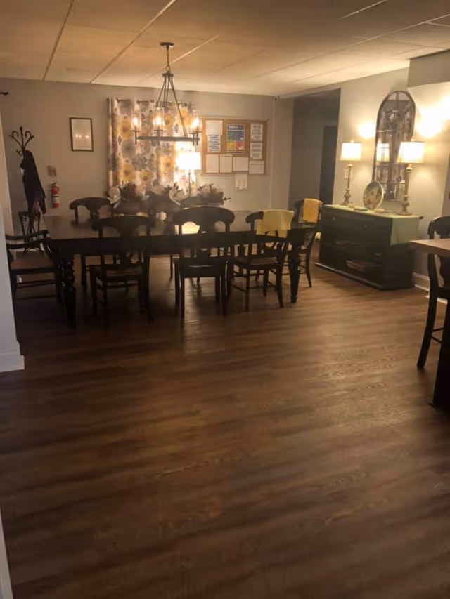 A warmly lit dining room with a long wooden table surrounded by chairs. The room features a chandelier above the table, floral curtains on the window, a coat rack with a jacket hanging, and a sideboard with two lamps and decorative items. The floor is wooden, and there are bulletin boards on the wall in the background.