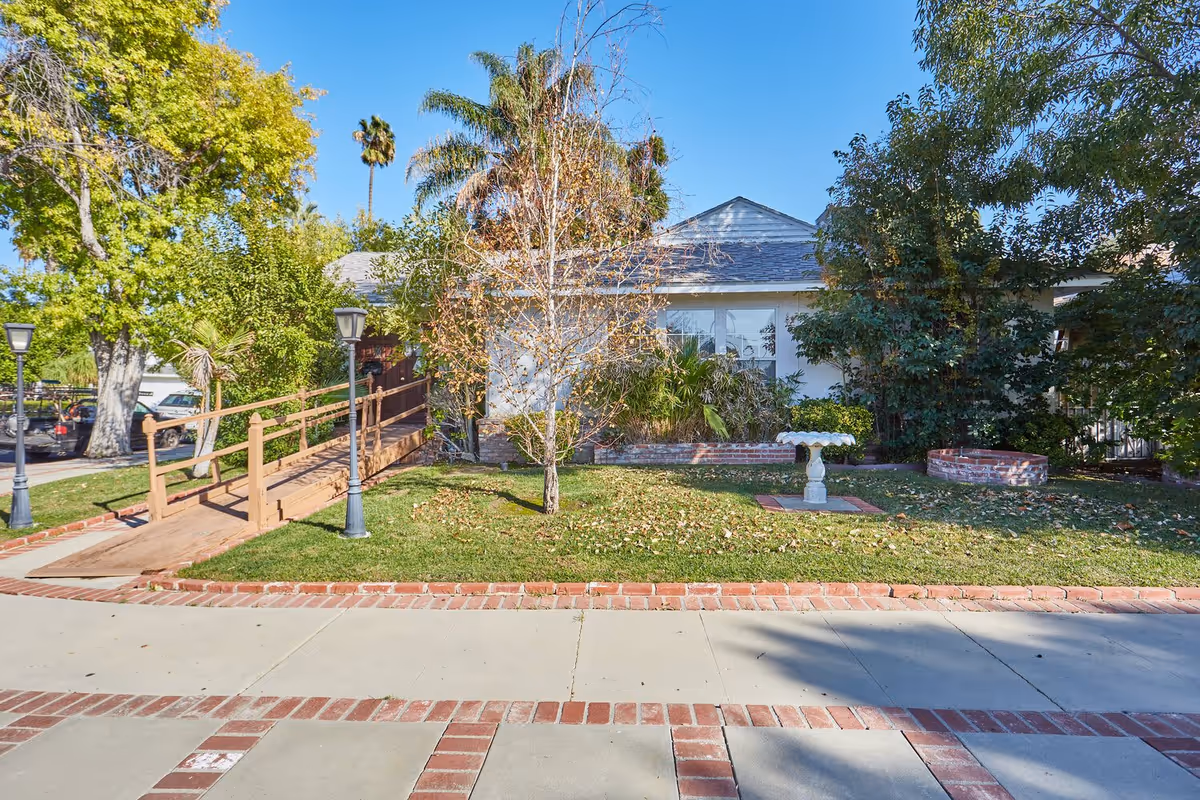 Front exterior of a single-story senior living building with a wooden wheelchair ramp, lawn, trees, and a birdbath.