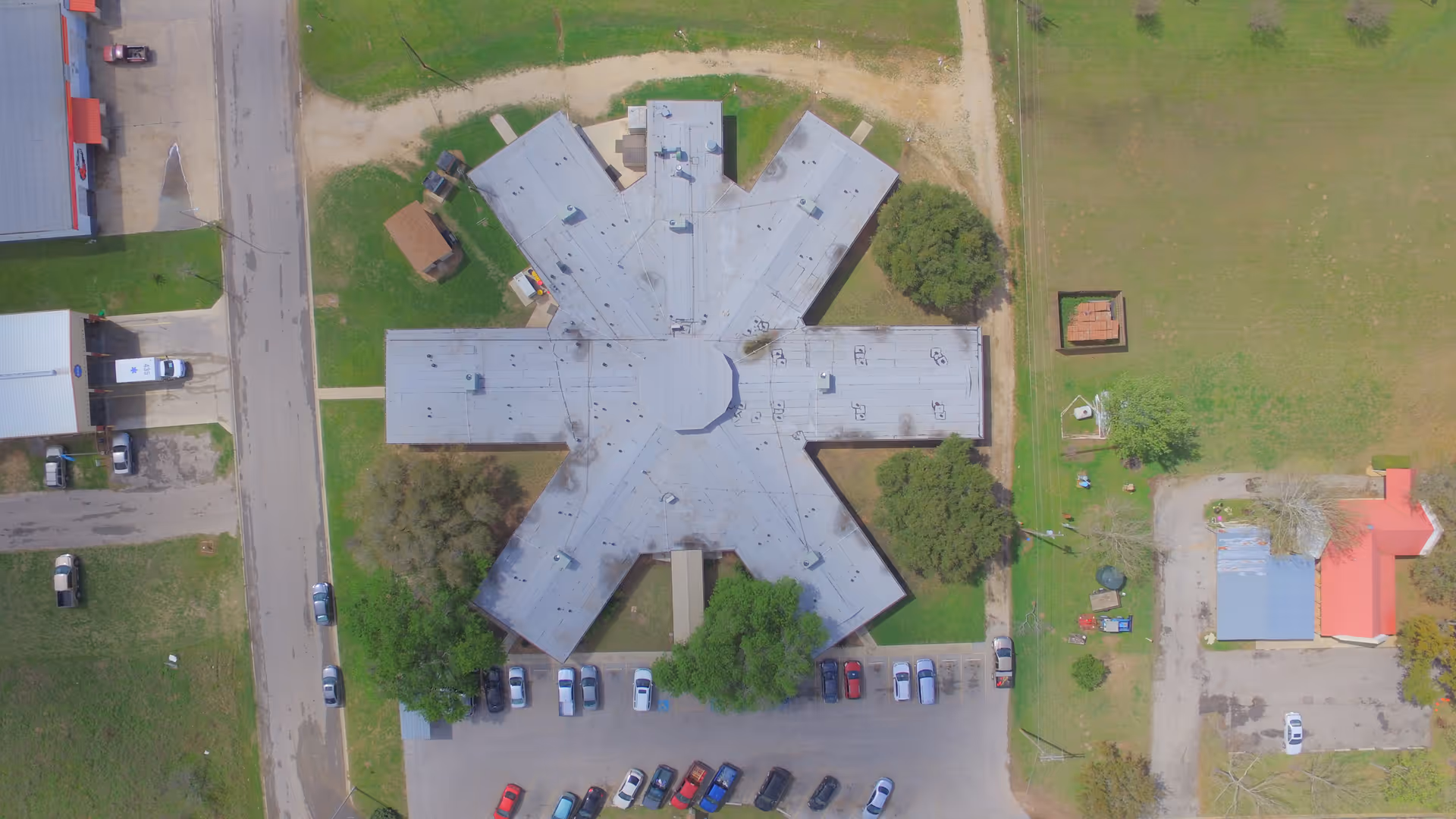 Aerial view of a star-shaped building with a parking lot in front, surrounded by grass, trees, and nearby smaller buildings and roads.
