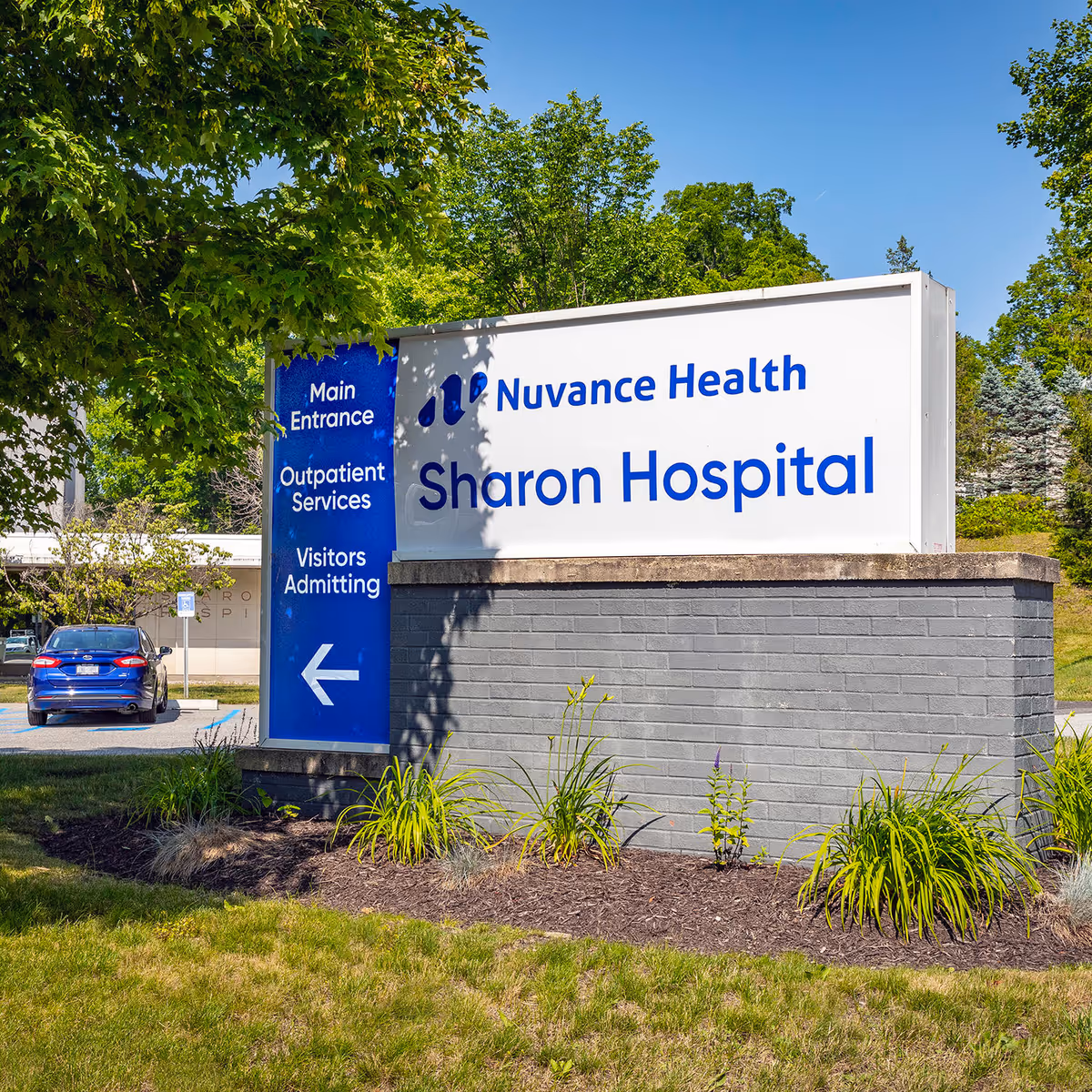 Exterior hospital sign reading Nuvance Health Sharon Hospital with greenery and a parked car nearby
