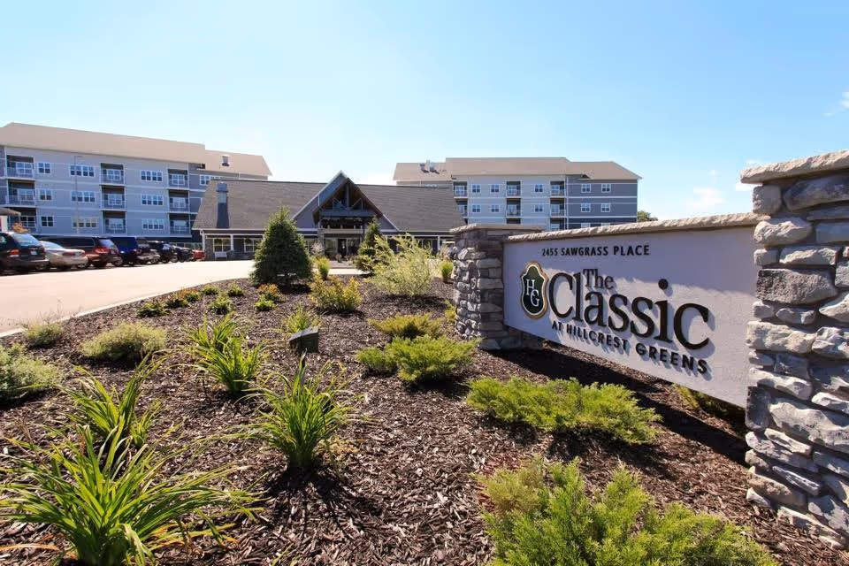 Exterior view of The Classic at Hillcrest Greens senior living facility showing a landscaped garden with various green plants and shrubs in the foreground, a stone sign with the facility name and address, a parking lot with cars, and a multi-story building with balconies in the background under a clear blue sky.
