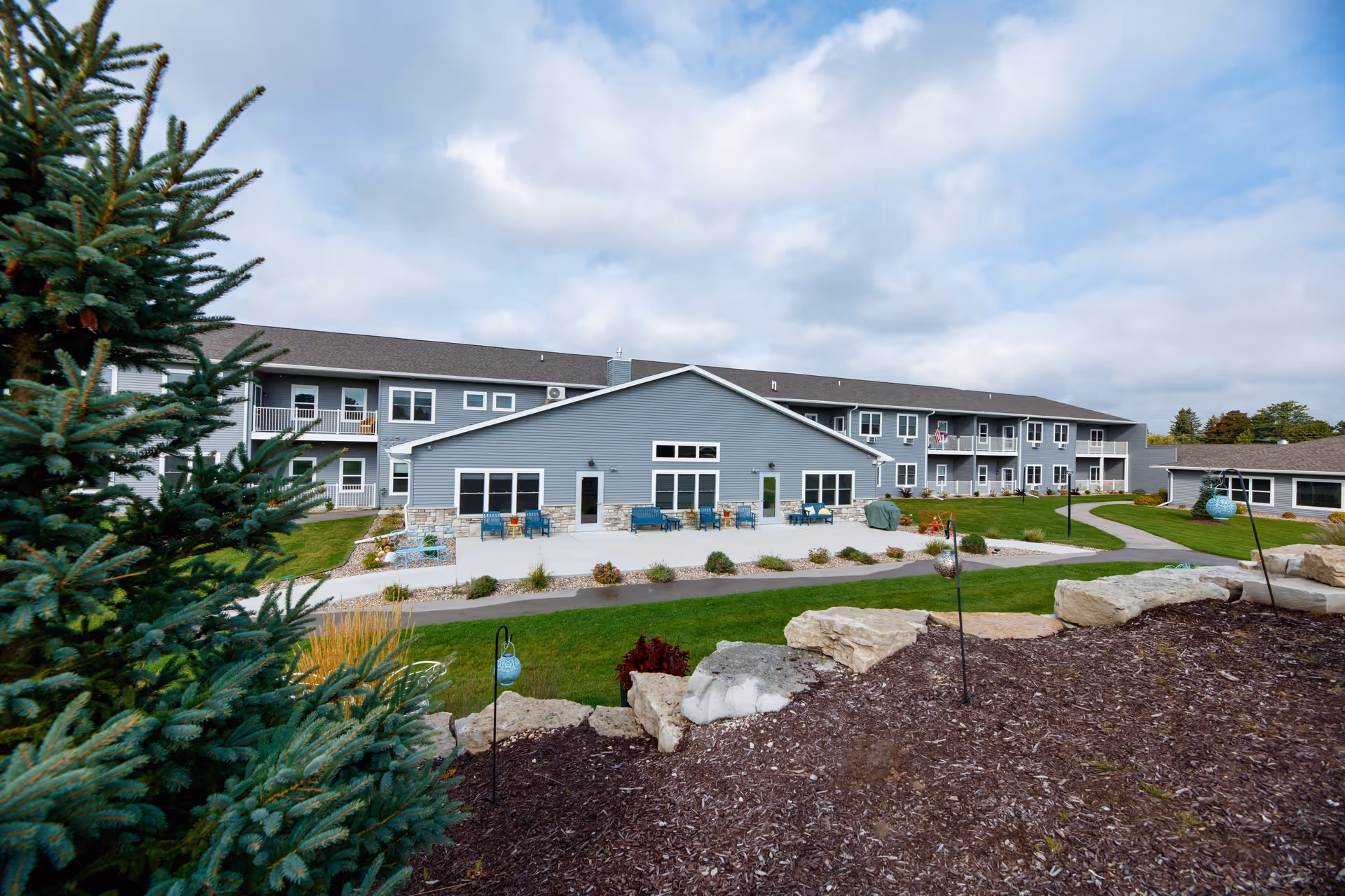 Exterior view of a two-story gray senior living building with patios, balconies, landscaped lawn and rock garden under a cloudy sky.