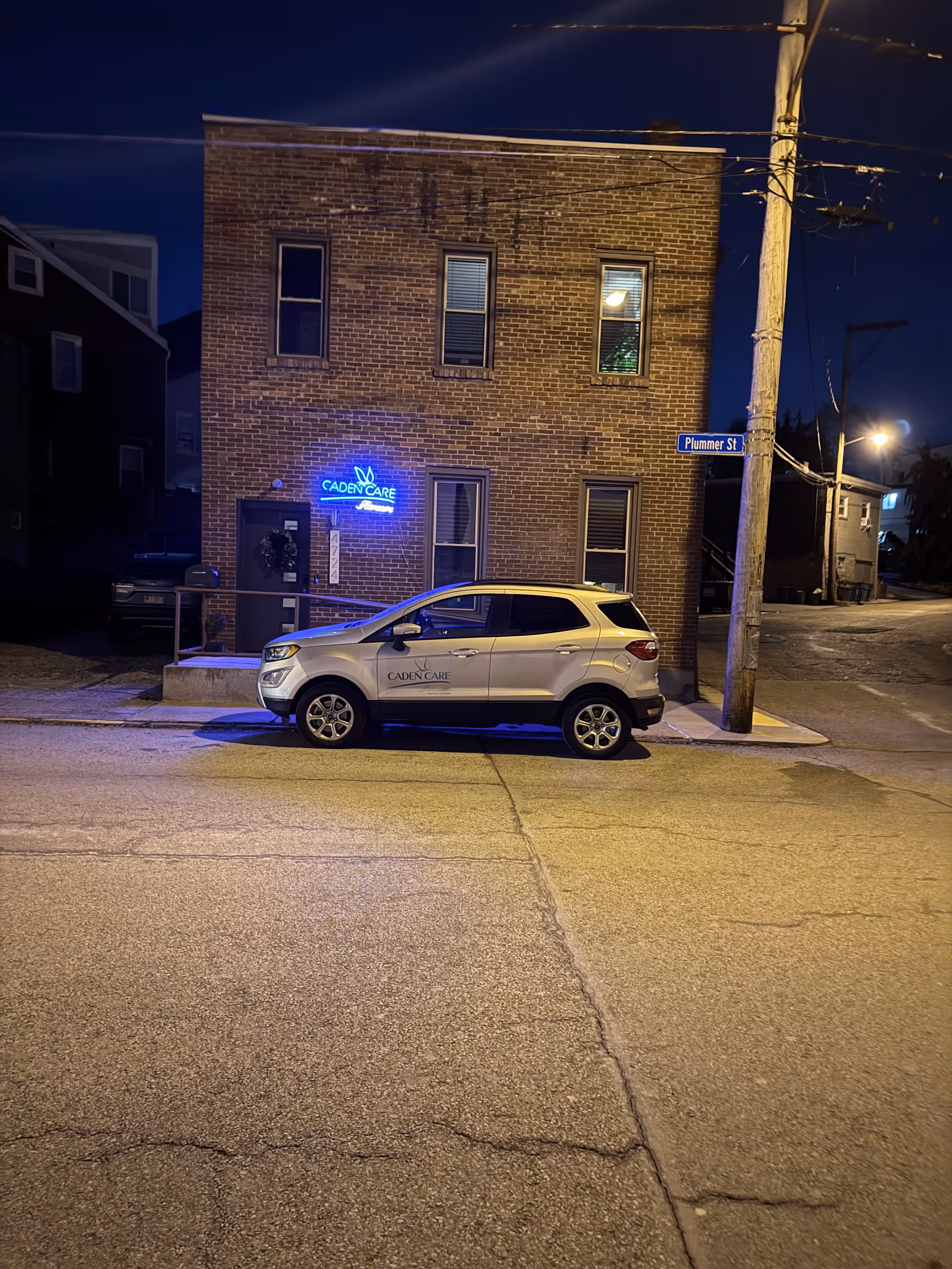 Night exterior of a brick building with blue neon sign and a parked Caden Care car.