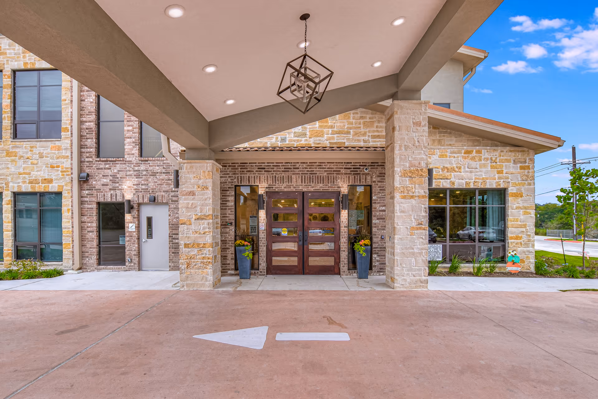 Covered porte-cochere entrance of a stone-and-brick senior living building with double wooden doors and potted plants.