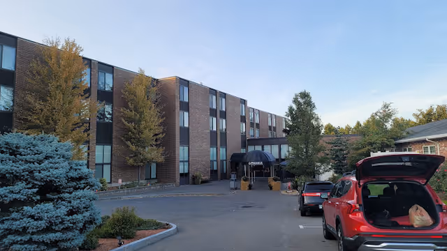 Front exterior of a three-story brick senior living facility with a covered entrance, parked cars, and landscaping.