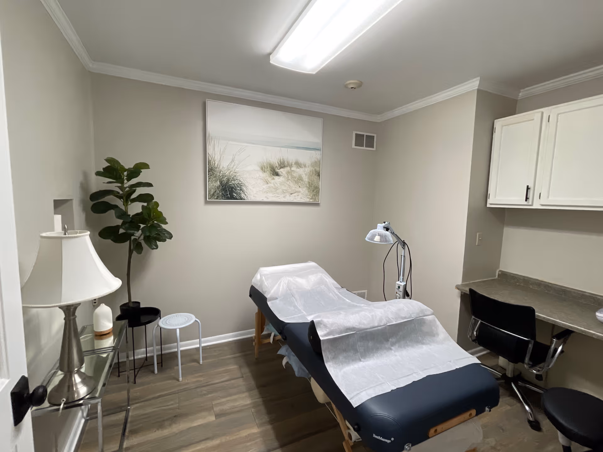 A small clinical treatment room with an adjustable chair, lamp, cabinets, and plant.