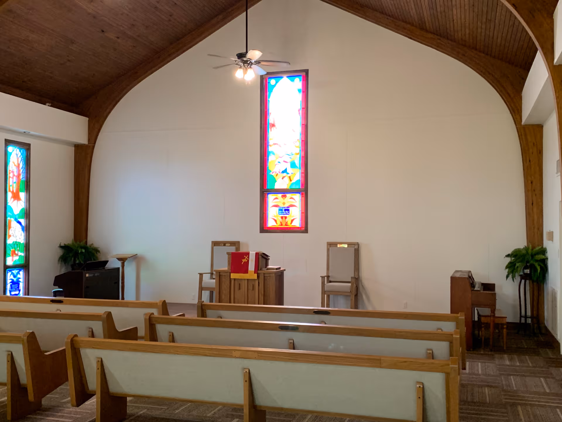 Interior view of a small chapel or worship room with wooden pews, a wooden arched ceiling, two stained glass windows, a ceiling fan with lights, a pulpit with a red cloth, two chairs, potted plants, and a small piano on the right side.
