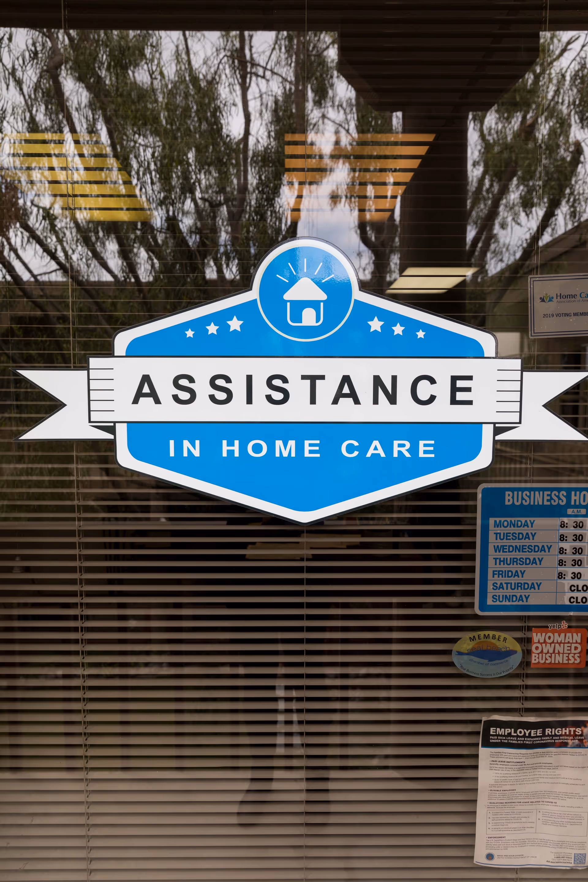 A blue storefront sign reads 'ASSISTANCE IN HOME CARE' on glass doors with blinds.