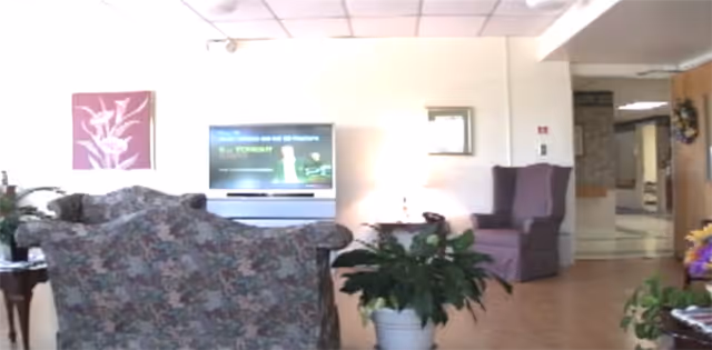 Living room in a senior facility with floral chairs, a TV, and plants.
