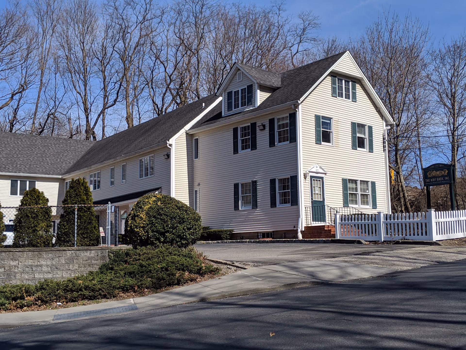 Two-story beige senior living facility with pitched roof, white fence, and surrounding trees.