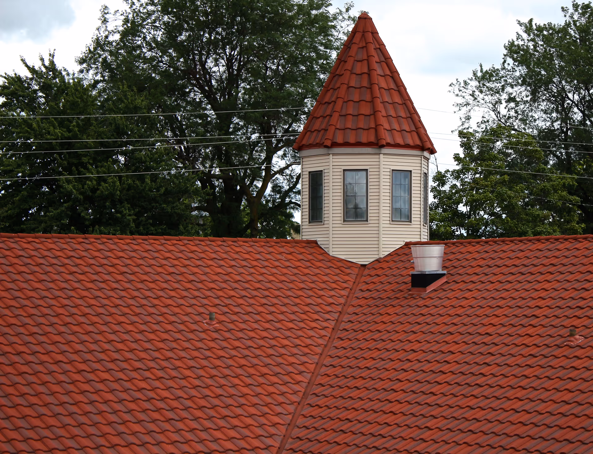 View of a building's red tiled roof with a small tower-like structure featuring windows and a pointed red roof, surrounded by green trees and power lines in the background.