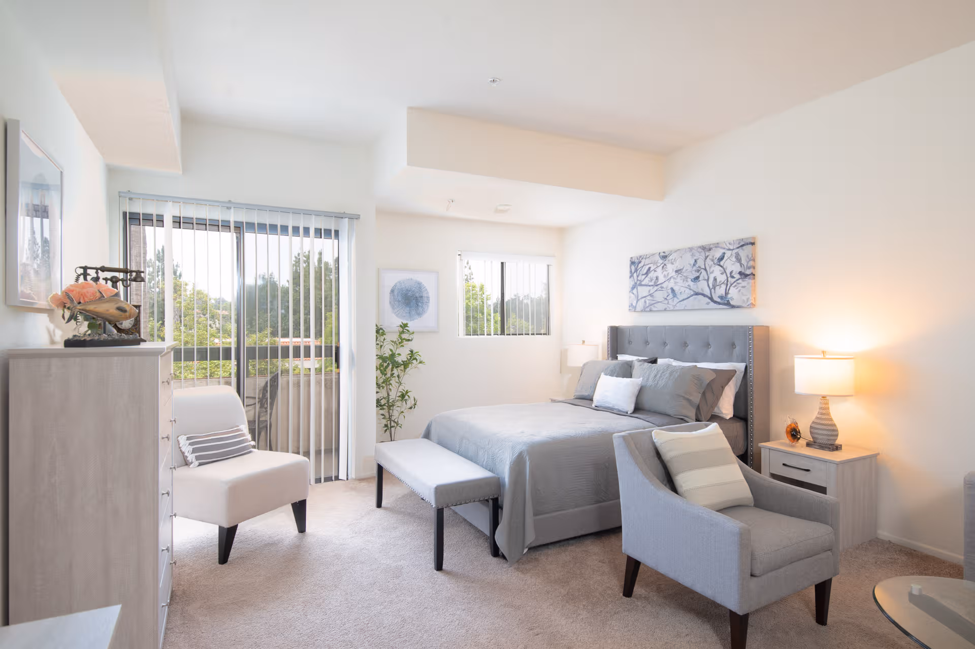 Bright, modern bedroom in a senior living facility with a gray bed, seating, and balcony.