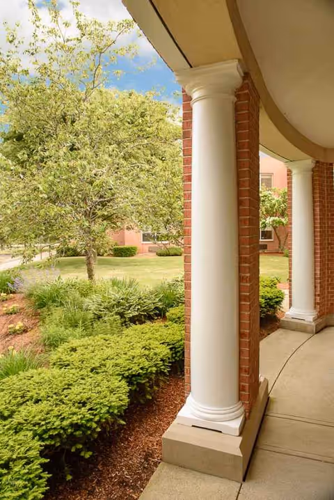 Covered walkway with white columns along a brick building and well-kept shrubs outside.